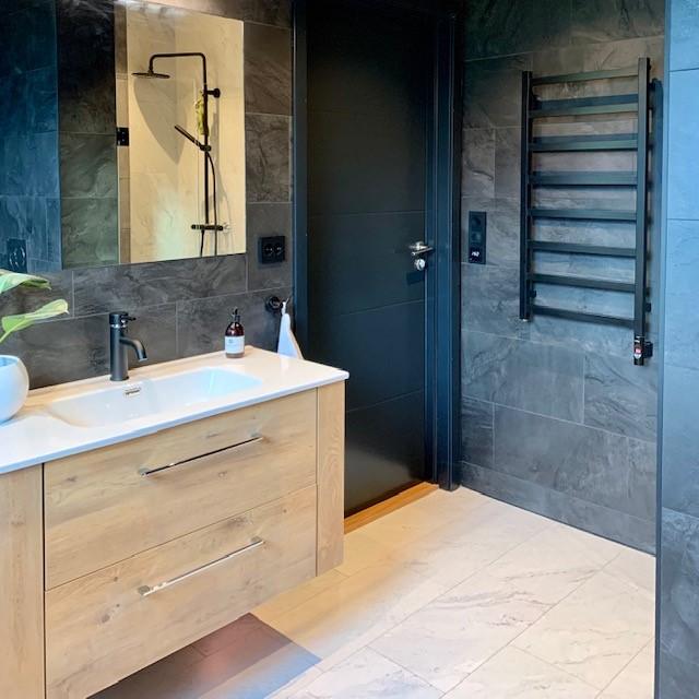 A finished bathroom with dark tiles, a wooden vanity and a heated towel rail