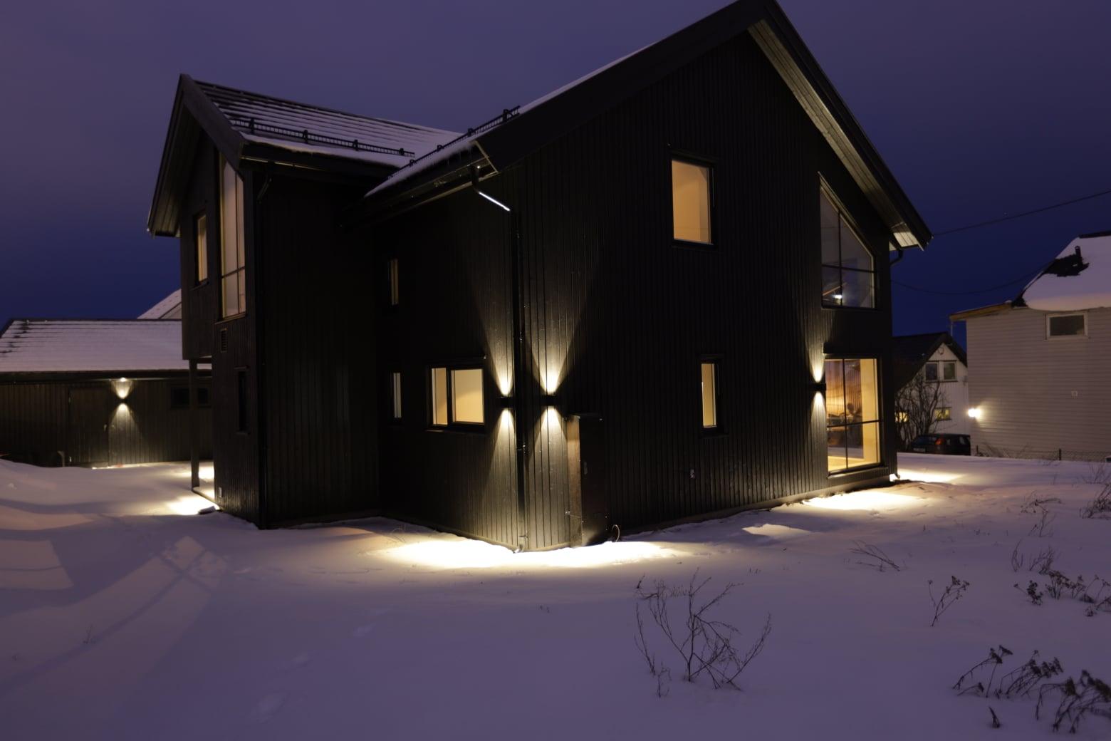 A dark-clad new house in the snow at dusk, lit from inside and by wall lights